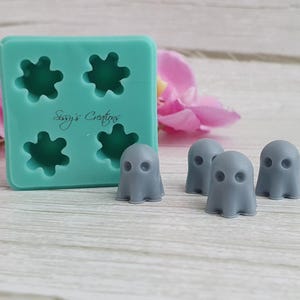 Puede incluir: Un molde de silicona turquesa con cuatro cavidades en forma de estrella y el texto "Sissy's Creations". Junto al molde, cuatro pequeñas figuras grises con forma de fantasma. El fondo incluye flores rosas y una superficie de madera clara.
