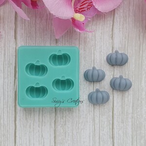 May include: A teal silicone mold with four pumpkin-shaped cavities and five gray pumpkin-shaped candies. The mold has the text "Sissy's Creations" on the bottom. The background includes pink and white flowers and a light wood-grain surface.