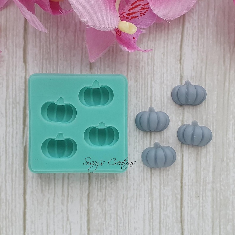May include: A teal silicone mold with four pumpkin-shaped cavities and five gray pumpkin-shaped candies. The mold has the text "Sissy's Creations" on the bottom. The background includes pink and white flowers and a light wood-grain surface.