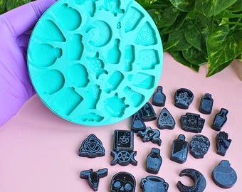 May include: A round, teal silicone mold featuring various recessed shapes including skulls, potion bottles, moons, and occult symbols. Beside the mold are several small, black, finished resin pieces that match the shapes within the mold.