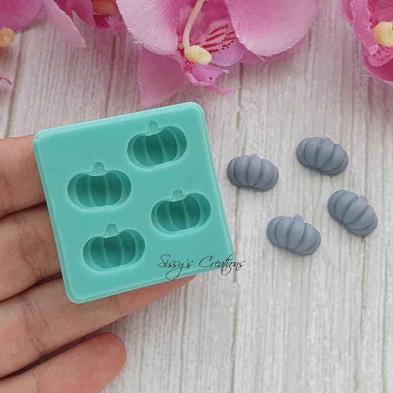 May include: A teal silicone mold with five pumpkin-shaped cavities and several small gray pumpkin-shaped decorations. The mold has the text "Sissy's Creations" on the bottom right. The pumpkins are for crafting or decoration.