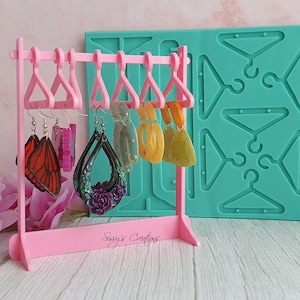 Puede incluir: Un expositor de pendientes rosa con cinco pares de pendientes colgados. Los pendientes están hechos de resina y presentan varios diseños, incluyendo mariposas, flores y formas geométricas. Un molde de silicona turquesa con múltiples formas de ganchos para pendientes se encuentra en el fondo. El texto "Sugie's Crafts" está impreso en el expositor.