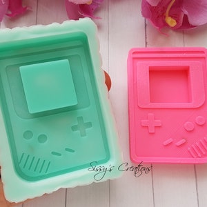 Può includere: Due stampi in silicone per realizzare oggetti in resina. Gli stampi hanno la forma di una console per videogiochi portatile retrò. Uno stampo è verde e l'altro è rosa.