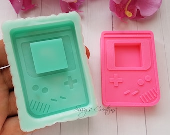 Puede incluir: Dos moldes de silicona para hacer manualidades de resina. Los moldes tienen forma de consola de videojuegos portátil retro. Un molde es verde y el otro es rosa.