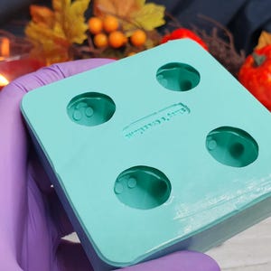 May include: A turquoise silicone mold with four round cavities. The mold is designed for making small round objects, such as candles or soap.