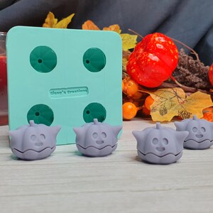May include: A teal silicone mold with four pumpkin-shaped cavities. Four gray pumpkin-shaped silicone molds are sitting on a wooden surface. The mold is labeled "Always Creating".