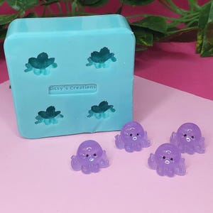 May include: A light blue silicone mould with four teal flower-shaped impressions and the text "Sissy's Creations". Four small, translucent purple octopus-shaped resin charms are displayed on a pink surface.