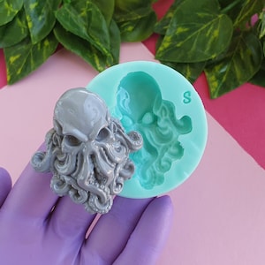 Pode incluir: Um molde de silicone cinza para fazer uma escultura de resina de um crânio de polvo. O molde é redondo e tem uma borda elevada. O molde é mostrado ao lado de uma escultura de resina cinza de um crânio de polvo.