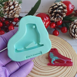 Peut inclure: Un moule en silicone bleu clair en forme de patins à glace avec un trou en haut. Le moule est tenu dans une main gantée. Un charme de patins à glace rouge est posé sur une surface en bois.