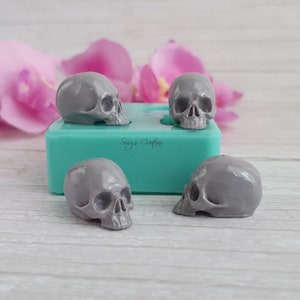Puede incluir: Un molde de silicona turquesa con el texto Sissy's Creations en relieve en la parte frontal, acompañado de tres pequeñas piezas de resina gris con forma de calavera, lisas, colocadas alrededor del molde sobre una superficie de madera clara.