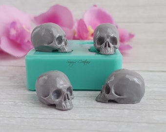 Op de afbeelding: Een turquoise siliconen mal met de tekst Sissy's Creations in reliëf op de voorzijde, vergezeld van drie kleine, gladde, grijze schedelvormige harsstukjes die rond de mal op een lichtgekleurd houten oppervlak zijn geplaatst.