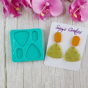 May include: A turquoise silicone mold with four shapes: two triangles, a rectangle, and an oval. Next to the mold are two pairs of earrings, one pair is green with gold flecks and orange, the other is green with gold flecks and orange. The card reads "Sizzy's Creations".