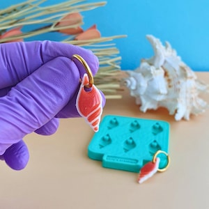 Puede incluir: Pendientes en forma de concha con aros dorados. Los pendientes son rojos y blancos, con un acabado brillante. Un molde azul y una concha marina están en el fondo. Los pendientes son sostenidos por una persona con guantes morados.