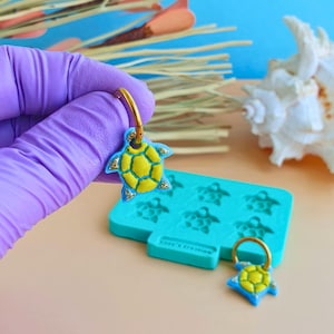Pode incluir: Brincos de tartaruga feitos à mão com argolas douradas. As tartarugas são amarelas com contornos azuis e detalhes em glitter dourado. No fundo, um molde de silicone azul com formas de tartaruga.