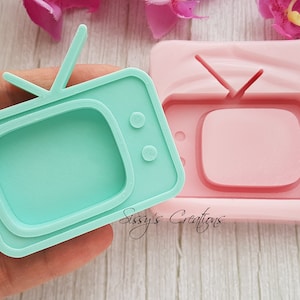 Pode incluir: Um molde de silicone para fazer artesanato de resina em forma de televisão vintage. O molde é rosa e tem uma televisão verde claro dentro.