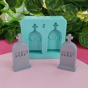 Puede incluir: Un molde de silicona para hacer dos pequeñas decoraciones con forma de lápida. El molde es azul claro y tiene dos cavidades con forma de lápida con una cruz en la parte superior y la palabra "RIP" inscrita en la parte delantera. Se muestran dos lápidas grises junto al molde.