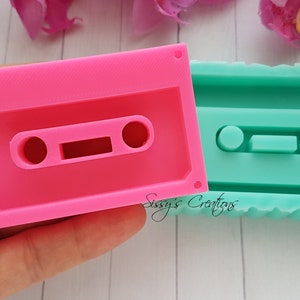 Può includere: Due stampi in silicone rosa e verde acqua a forma di cassette. Gli stampi sono progettati per creare piccole repliche decorative di cassette.