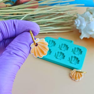May include: A pair of seashell-shaped earrings with gold-colored hoops. The earrings are orange and white with glitter accents. A turquoise mold with seashell shapes is in the background.