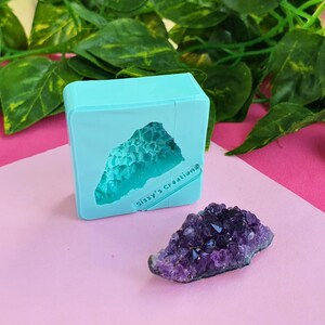 Peut inclure: Un moule en silicone bleu clair de forme carrée présentant une cavité en creux aux bords irréguliers rappelant un cristal, avec le texte Sissy's Creations en relief, à côté d'un amas de cristaux d'améthyste violet foncé aux facettes pointues.