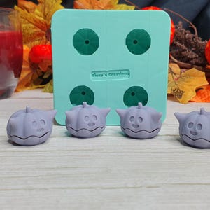 May include: Four small purple pumpkin-shaped silicone molds with a cute face design. The molds are sitting on a light wood surface next to a green silicone mold with four cavities.