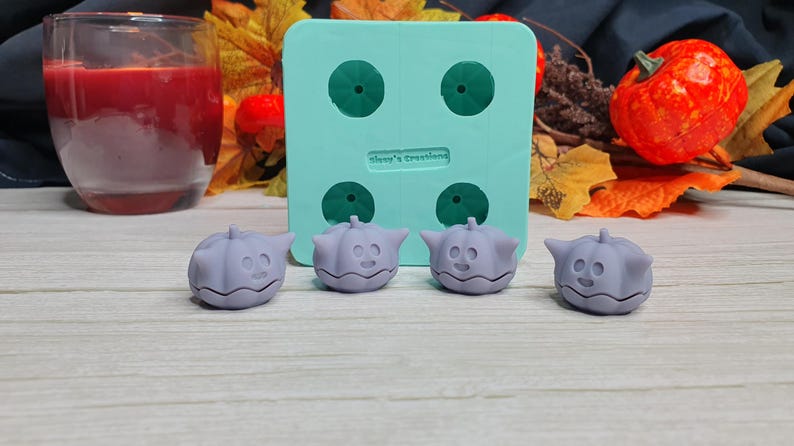 May include: Four small purple pumpkin-shaped silicone molds with a cute face design. The molds are sitting on a light wood surface next to a green silicone mold with four cavities.