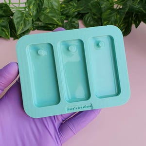 Può includere: Uno stampo in silicone verde acqua chiaro con tre cavità rettangolari, ciascuna con una piccola sporgenza circolare. Lo stampo è rettangolare con angoli arrotondati e il testo "Sissy's Creations" è stampato in basso. Lo stampo è tenuto da una mano con un guanto viola.