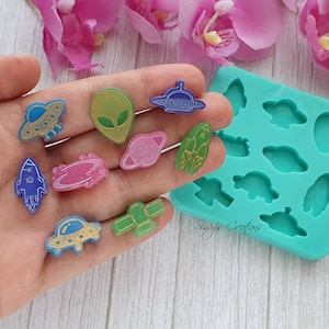 May include: A hand displays several colourful resin charms shaped like UFOs, aliens, rockets, and planets. A teal silicone mould with similar shapes is next to the hand. The charms are in various colours, including blue, green, pink, and yellow.