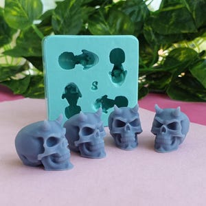 Puede incluir: Una colección de cuatro figuritas de calaveras de diablo grises con cuernos, junto a un molde de silicona turquesa. El molde presenta cavidades con forma de calaveras y otras figuras. Las calaveras están sobre una superficie rosa con follaje verde de fondo.