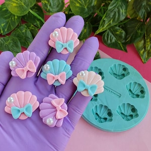 Könnte beinhalten: Eine Silikonform mit sechs Muschelformen, die zum Erstellen von Harzschmuck verwendet wird. Die Form ist türkisblau und die Muscheln sind rosa, lila und blau. Es gibt sechs fertige Muscheln mit Perlenakzenten und Schleifen.