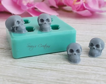 May include: A rectangular teal silicone mold with multiple cavities for casting small skull shapes. Two grey skull figures are positioned on top of the mold, and two additional grey skull figures sit beside it. The text Sissy's Creations is printed on the mold.