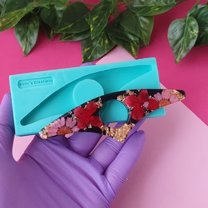 Può includere: Uno stampo in silicone per realizzare anelli in resina. Lo stampo è di colore blu turchese e ha una singola forma ad anello. Un anello in resina finito con una base nera, dettagli dorati e fiori pressati rossi e rosa è mostrato accanto allo stampo.