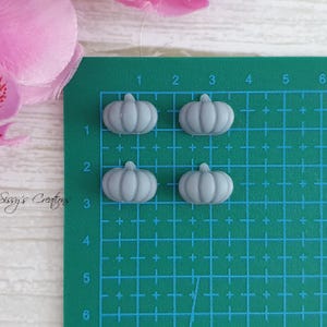 May include: Four small, light gray pumpkin-shaped embellishments arranged on a green cutting mat. The pumpkins have a ribbed texture and a small stem. A pink flower is visible in the upper left corner.