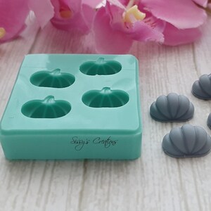 May include: A teal silicone mold with four pumpkin shapes and the text "Sissy's Creations". Several gray pumpkin-shaped objects are next to the mold. The background includes pink and white flowers.