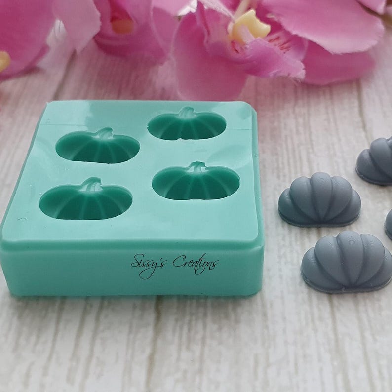 May include: A teal silicone mold with four pumpkin shapes and the text "Sissy's Creations". Several gray pumpkin-shaped objects are next to the mold. The background includes pink and white flowers.