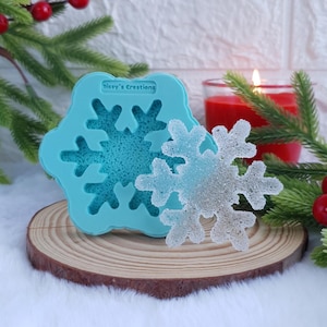 Può includere: Uno stampo per fiocchi di neve in silicone blu chiaro con il testo "Sissy's Creations" e un fiocco di neve trasparente e glitterato. Lo stampo e il fiocco di neve sono su una fetta di legno, con verde e una candela rossa sullo sfondo. Tema festivo.