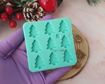 May include: A teal silicone mould with nine Christmas tree shapes. The mould is held in a purple glove. The mould is on a wooden surface with gold glitter ornaments.