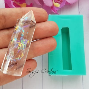 以下が含まれることがあります： 虹色のグリッターが埋め込まれた透明なクリスタル型のモールド。モールドは手で持たれており、隣にはターコイズブルーのシリコンモールドがあります。シリコンモールドには、長方形のくぼみが1つあります。