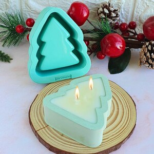 May include: A teal Christmas tree-shaped candle and mold sit on a wooden surface, with festive decorations in the background. The candle has two lit wicks, and the mold is empty. The candle and mold are both teal.