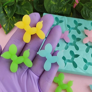 Op de afbeelding: Een siliconen mal voor het maken van ballonhondvormige harscharms. De mal is turquoiseblauw en heeft 16 holtes. Er zijn vier afgewerkte charms in verschillende kleuren: geel, groen, blauw en roze.