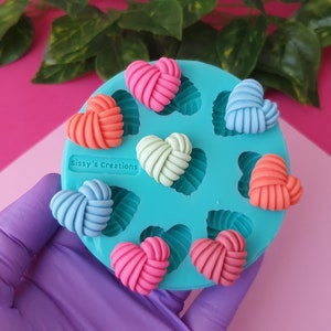 Puede incluir: Un molde de silicona turquesa con ocho cavidades en forma de corazón. El molde está lleno de piezas de resina en forma de corazón anudadas y de colores. El molde está etiquetado como "Sissy's Creations".