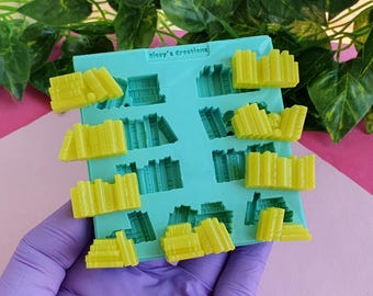 Könnte beinhalten: Eine hellblaue Silikonform mit acht Vertiefungen in Form von Bücherstapeln. Der Text Sissy's Creations ist oben eingeprägt. Mehrere leuchtend gelbe Bücherstapel-Formen aus Harz liegen in den Vertiefungen der Form.
