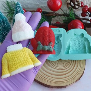 May include: A collection of Christmas-themed silicone molds and finished products. Includes a pink and white knit hat, a yellow sweater, and a red mitten with a green bow. A blue mold with a sweater design is also visible.