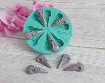 May include: A teal silicone mold with eight gray bird skull impressions, alongside six cast skulls. The mold is labeled "Sissy's Creations". Pink orchids are in the background. The skulls are a detailed, gothic-themed craft supply.