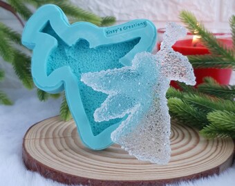 May include: A clear, sparkly angel figurine next to a light blue silicone mold. The mold has the text "Sissy's Creations" on it. The angel is on a wooden slice, with a red candle and greenery in the background.
