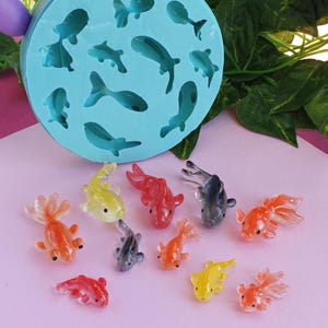 May include: A light blue silicone mould with fish shapes and several colourful resin goldfish. The goldfish are in shades of orange, yellow, red, and grey. The mould is round and sits on a pink surface.