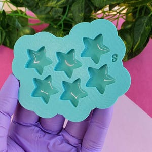 Puede incluir: Un molde de silicona azul claro con seis cavidades en forma de estrella dispuestas en grupo. El molde tiene un acabado exterior mate y liso con formas de estrella cóncavas y brillantes. La letra S está grabada cerca de la estrella superior derecha.