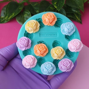 Może przedstawiać: Okrągła, turkusowa forma silikonowa z wieloma wgłębieniami w kształcie róż, z wytłoczonym na środku napisem Sissy's Creations. Na wierzchu umieszczono kilka gotowych elementów w kształcie róż w pastelowych odcieniach pomarańczu, żółci, błękitu, różu i fioletu.