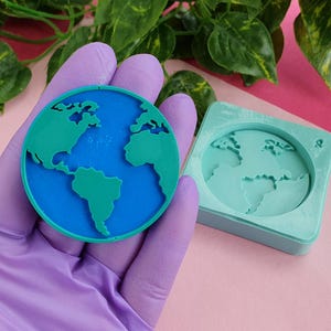 May include: A blue and green 3D printed earth globe resin mould. The mould is made of silicone and is flexible. The mould is in the shape of a circle and has a raised design of the earth. The mould is on a pink surface.