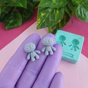 Peut inclure: Deux figurines grises, petites et de forme humaine, avec de grands yeux, réalisées à l'aide d'un moule en silicone. Le moule est bleu clair et porte l'inscription "Creations".