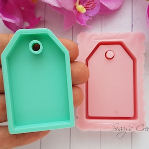 Puede incluir: Un molde de silicona verde para hacer etiquetas de resina. El molde tiene forma de etiqueta con un agujero en la parte superior. El molde se muestra junto a un molde de silicona rosa de la misma forma.